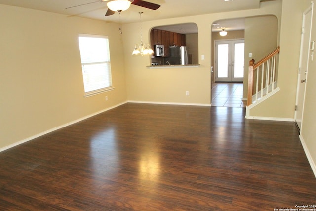 unfurnished living room featuring stairs, dark wood-type flooring, ceiling fan with notable chandelier, and baseboards