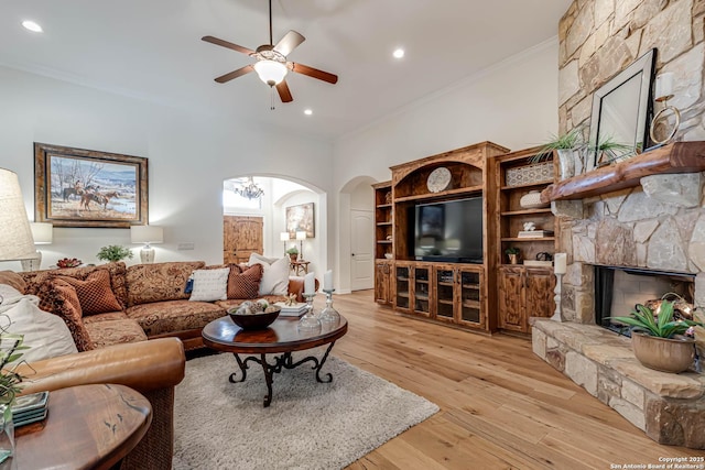 living area with crown molding, arched walkways, a ceiling fan, a stone fireplace, and light wood finished floors