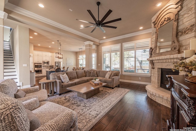 living area with ceiling fan, dark wood finished floors, ornamental molding, a stone fireplace, and stairs