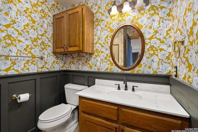 half bath with vanity, toilet, a wainscoted wall, and wallpapered walls