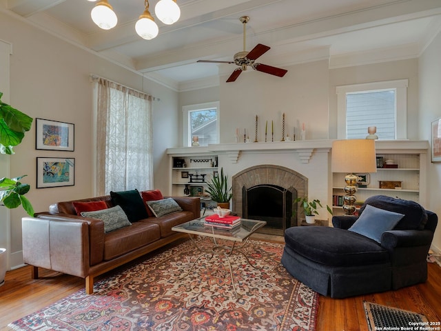 living room with a ceiling fan, beamed ceiling, a brick fireplace, and wood finished floors