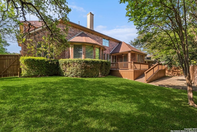 rear view of property featuring a chimney, brick siding, a wooden deck, fence, and a lawn