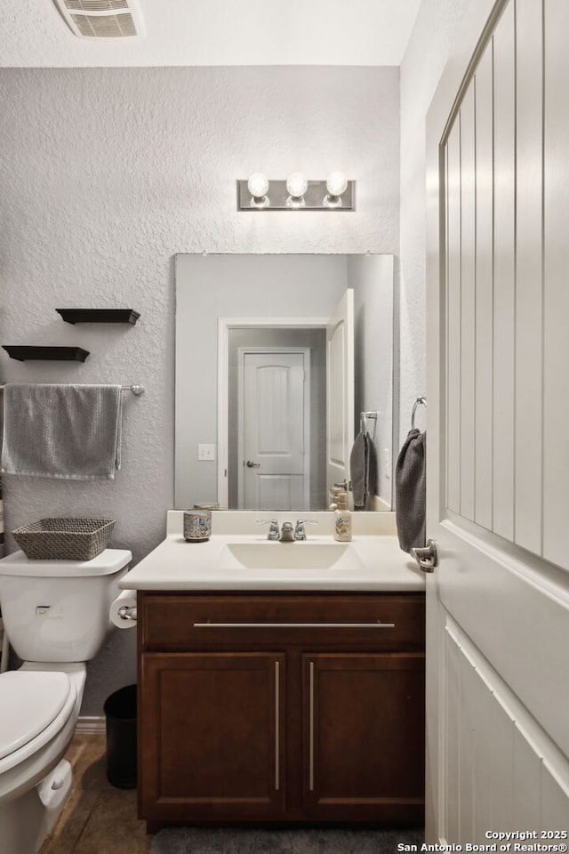 half bathroom with visible vents, toilet, vanity, and a textured wall