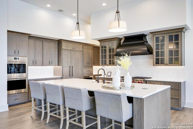kitchen featuring custom exhaust hood, light countertops, an island with sink, double oven, and light wood finished floors