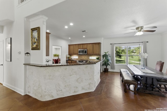 kitchen with brown cabinetry, tasteful backsplash, a ceiling fan, dark stone countertops, and stainless steel microwave