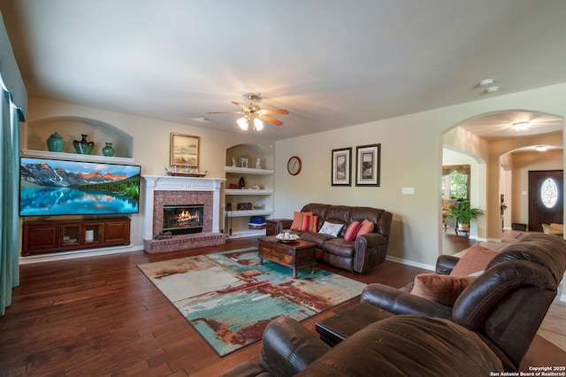 living area featuring built in features, arched walkways, a ceiling fan, a fireplace, and wood finished floors