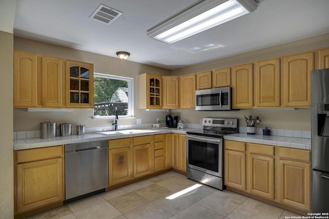 kitchen with light brown cabinets, glass insert cabinets, visible vents, appliances with stainless steel finishes, and a sink