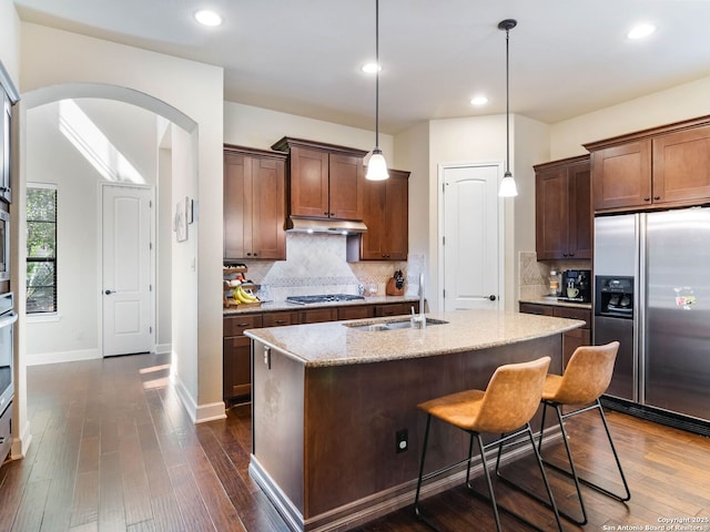 kitchen with arched walkways, appliances with stainless steel finishes, light stone counters, a sink, and under cabinet range hood
