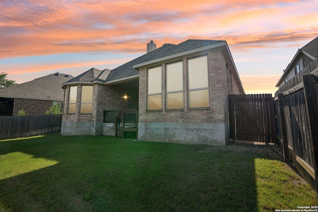 back of house at dusk featuring brick siding, a chimney, a fenced backyard, and a lawn