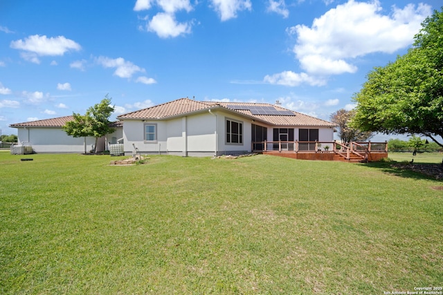 rear view of property featuring a wooden deck, a tiled roof, a sunroom, a lawn, and roof mounted solar panels