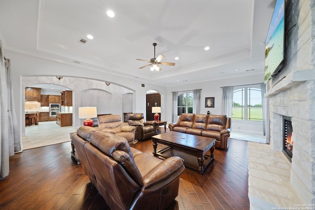 living area featuring arched walkways, a stone fireplace, a ceiling fan, dark wood finished floors, and a tray ceiling