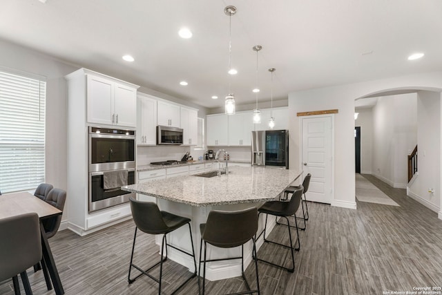 kitchen with baseboards, an island with sink, stainless steel appliances, arched walkways, and a sink