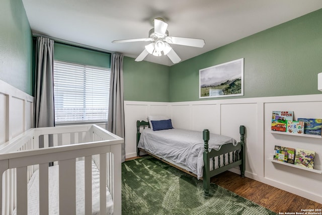 bedroom with a ceiling fan, wood finished floors, wainscoting, and a decorative wall