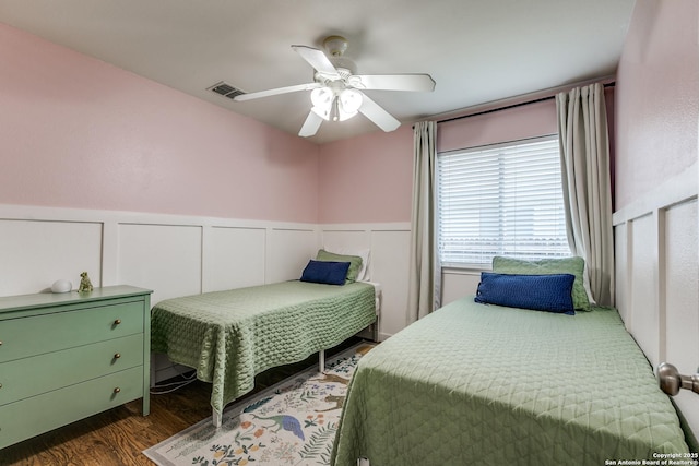 bedroom featuring visible vents, a decorative wall, ceiling fan, dark wood finished floors, and wainscoting
