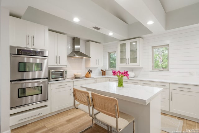 kitchen featuring white cabinets, stainless steel appliances, wall chimney range hood, glass insert cabinets, and tasteful backsplash