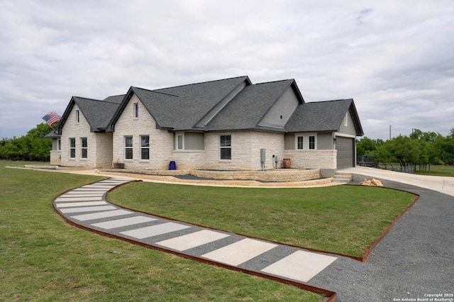 view of front facade featuring roof with shingles, a front yard, stone siding, an attached garage, and driveway