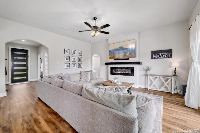 living area with a glass covered fireplace, arched walkways, a ceiling fan, and wood finished floors