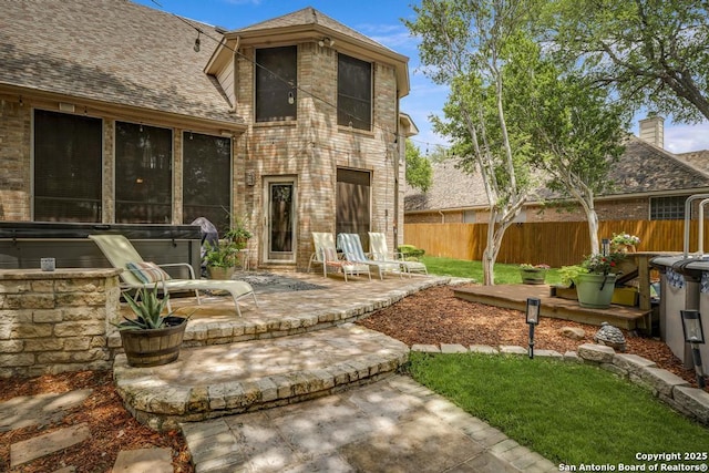 back of property featuring fence, roof with shingles, a hot tub, brick siding, and a patio area