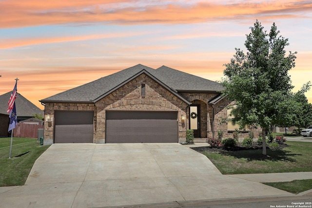 french country style house with roof with shingles, driveway, brick siding, an attached garage, and a yard