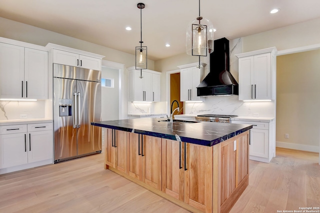 kitchen with wall chimney exhaust hood, a kitchen island with sink, light wood-style flooring, dark countertops, and stainless steel refrigerator with ice dispenser