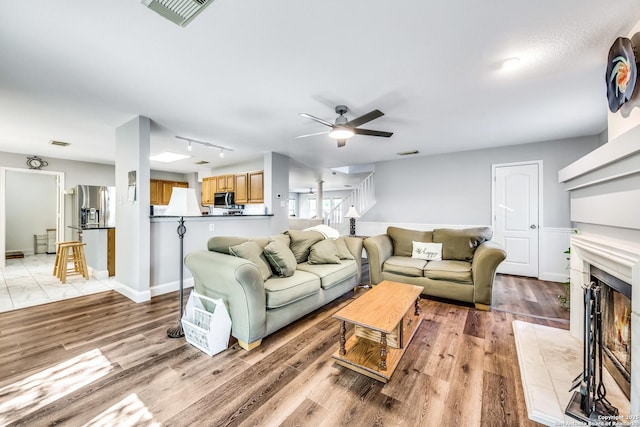 living room featuring stairway, visible vents, a warm lit fireplace, and ceiling fan