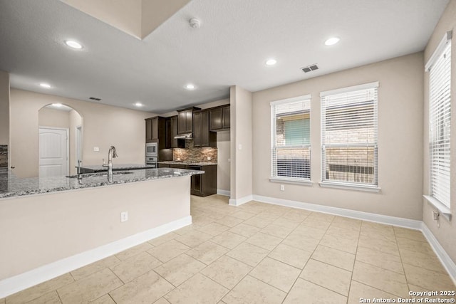 kitchen with arched walkways, a sink, visible vents, dark brown cabinetry, and light stone countertops