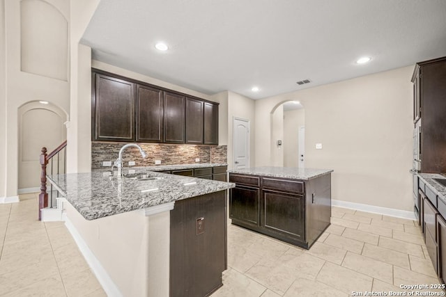 kitchen featuring arched walkways, a sink, dark brown cabinetry, decorative backsplash, and a kitchen island