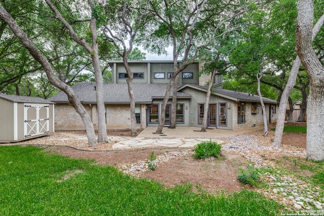 back of house featuring a storage shed, stone siding, a patio area, a shingled roof, and an outdoor structure