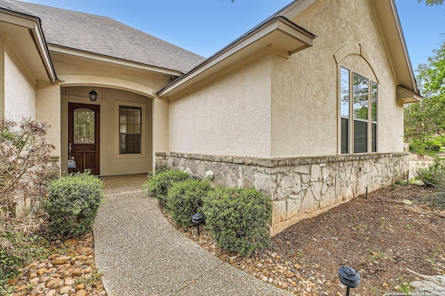 view of exterior entry with stucco siding and a shingled roof
