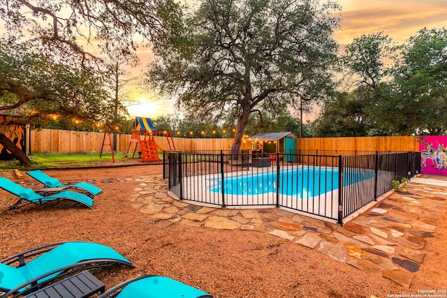 pool at dusk featuring a storage shed, a fenced in pool, a fenced backyard, a playground, and a patio