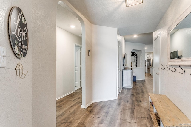 hall with wood finished floors, a textured wall, arched walkways, baseboards, and a textured ceiling