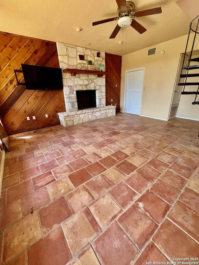 unfurnished living room with wooden walls, ceiling fan, a stone fireplace, visible vents, and a textured ceiling