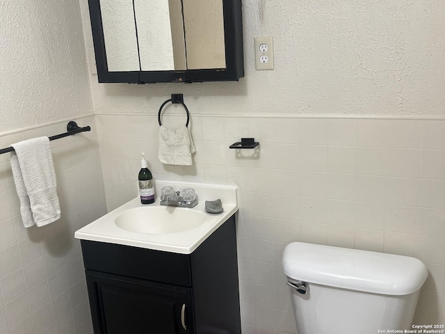 half bathroom featuring a wainscoted wall, a textured wall, toilet, vanity, and tile walls