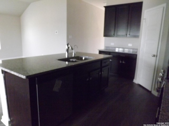 kitchen with a kitchen island with sink, a sink, dark wood finished floors, dark brown cabinets, and dark countertops