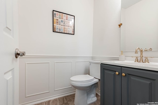 half bath featuring vanity, wainscoting, toilet, and a decorative wall