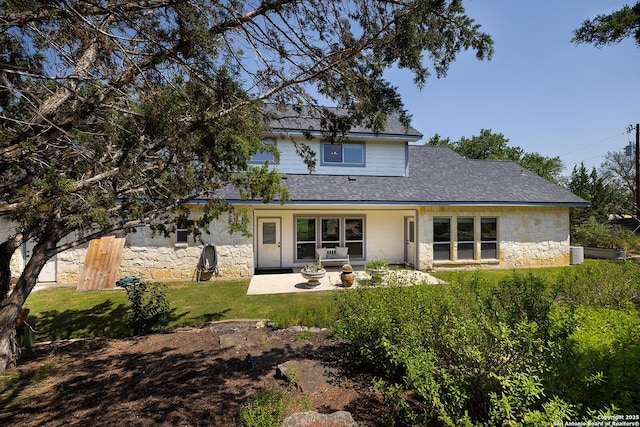 rear view of house featuring a patio area, stone siding, a shingled roof, and a lawn