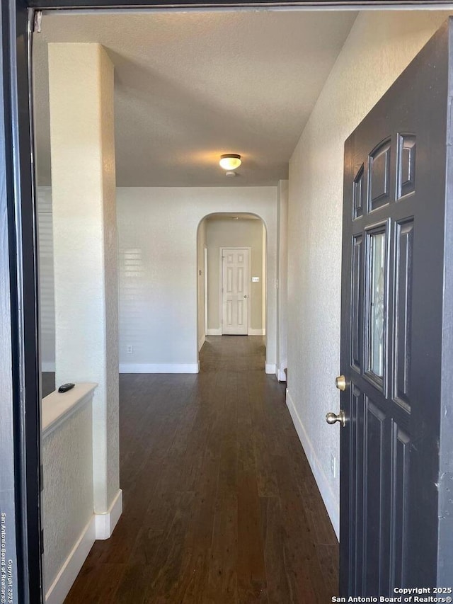 hall featuring a textured wall, baseboards, dark wood-style floors, and arched walkways