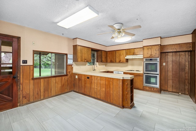 kitchen featuring a peninsula, light countertops, brown cabinetry, and stainless steel double oven