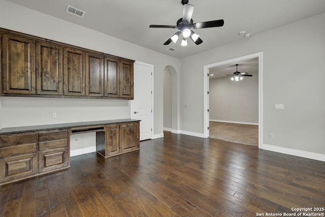 unfurnished office with built in study area, ceiling fan, arched walkways, dark wood-type flooring, and visible vents