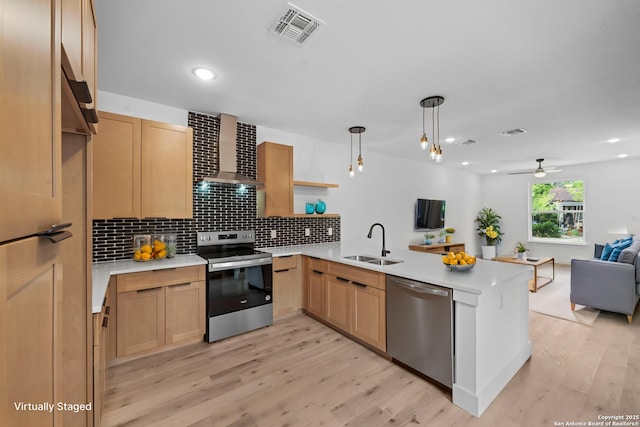 kitchen featuring wall chimney range hood, a sink, a peninsula, appliances with stainless steel finishes, and visible vents