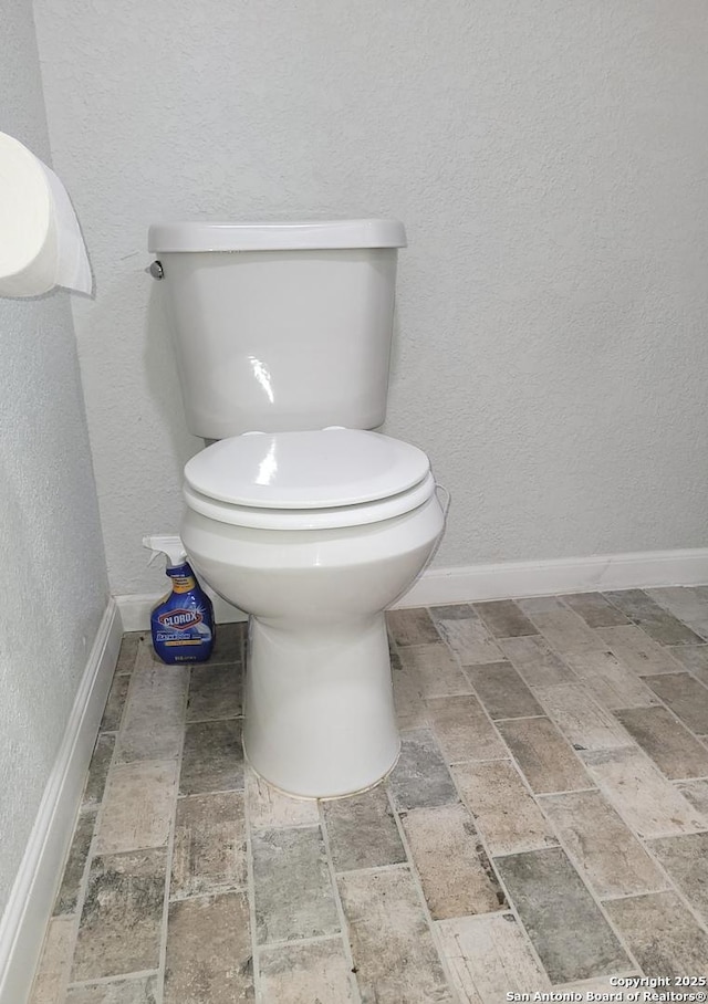bathroom featuring a textured wall, toilet, brick floor, and baseboards