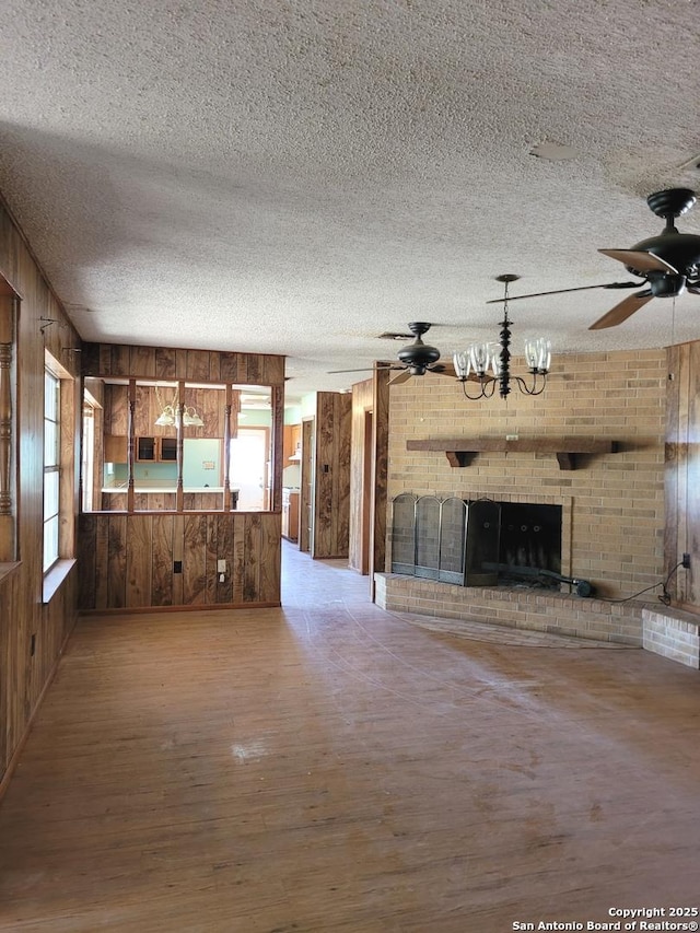 unfurnished living room featuring wood finished floors, a textured ceiling, ceiling fan with notable chandelier, wood walls, and a brick fireplace