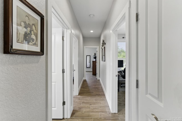 hallway featuring baseboards, a textured wall, light wood-style floors, and recessed lighting