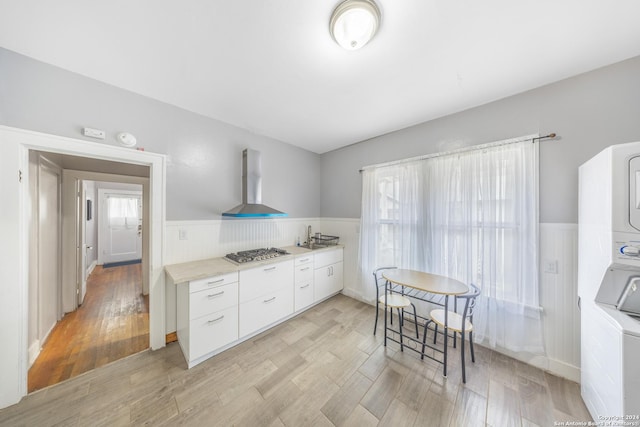kitchen with wall chimney range hood, stainless steel gas stovetop, wainscoting, stacked washer / drying machine, and white cabinetry
