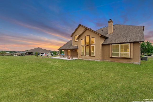 back of house at dusk with a lawn, a chimney, a shingled roof, and a patio area