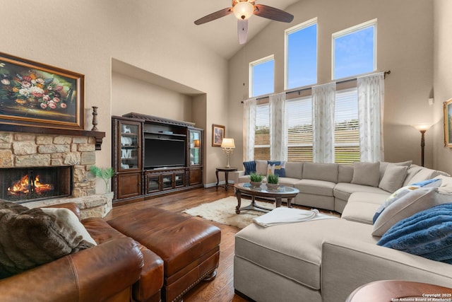 living room featuring high vaulted ceiling, a ceiling fan, a stone fireplace, baseboards, and wood finished floors