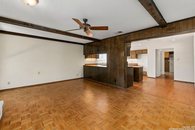 unfurnished living room with wood walls, a ceiling fan, beam ceiling, baseboards, and a textured ceiling