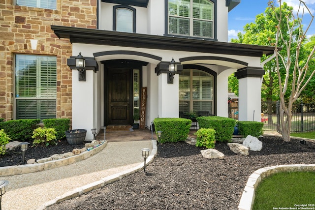 doorway to property with stucco siding, stone siding, covered porch, and fence