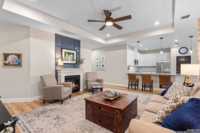 living room featuring light wood finished floors, baseboards, visible vents, a warm lit fireplace, and ceiling fan