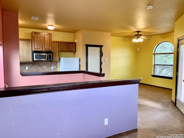 kitchen featuring brown cabinetry, stainless steel microwave, freestanding refrigerator, tasteful backsplash, and ceiling fan